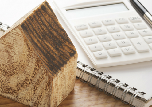 Small wooden carved house sitting on a desk with a a calculator and a noted pad with a book.