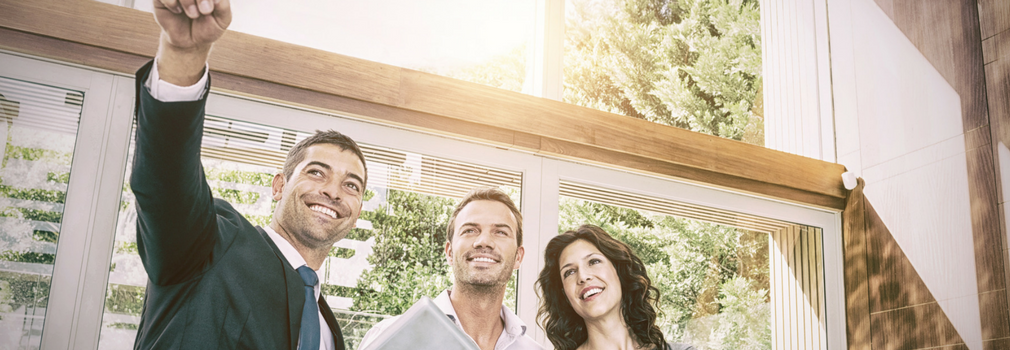 A real estate agent standing with two clients a man and a woman. Smiling and pointing to the house.